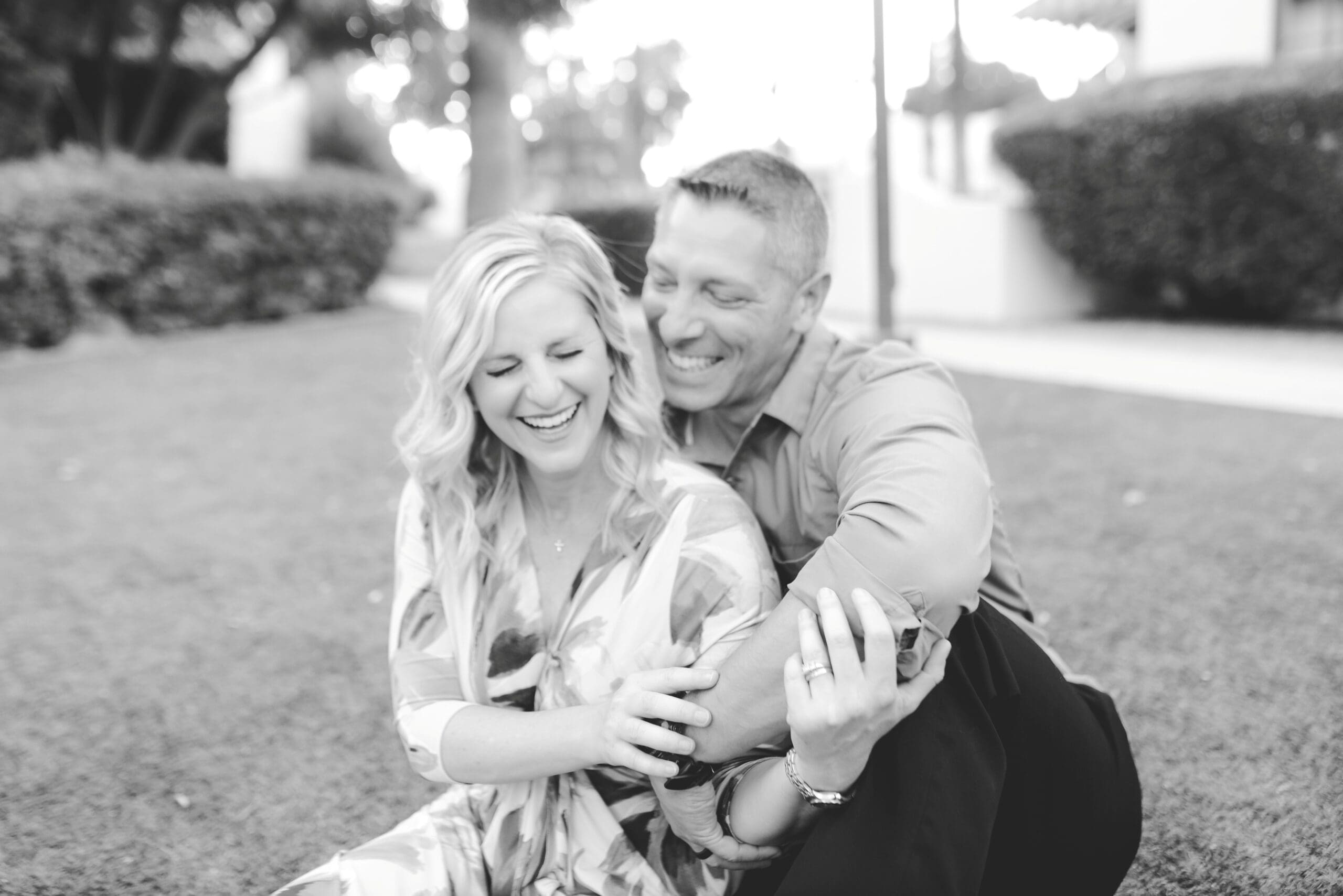 A joyful couple sits on the grass, laughing together in a heartfelt moment. The man wraps his arms around his wife as she playfully holds his arm, both radiating love and connection.