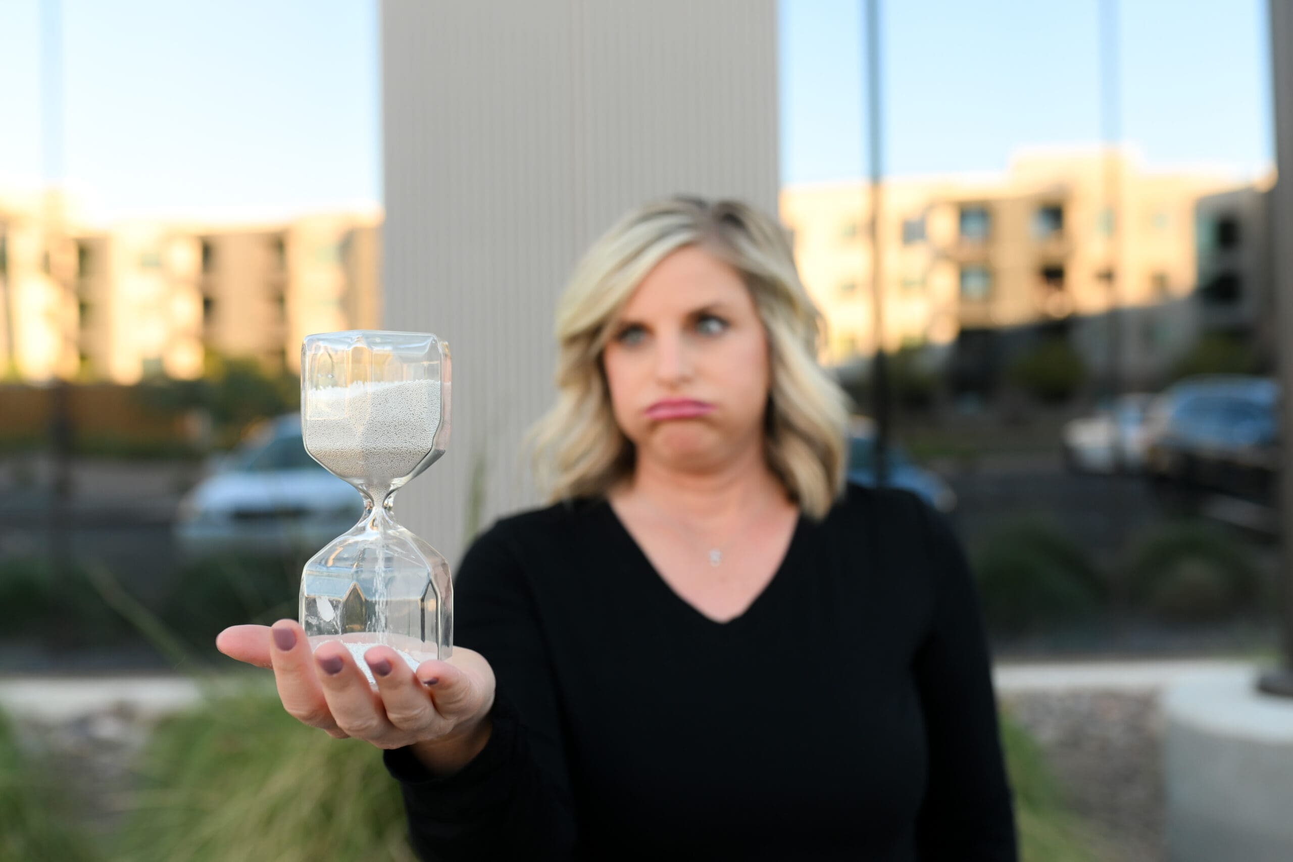 A blonde woman wearing a black shirt holds a clear hourglass with sand flowing through it while making a frustrated expression, symbolizing the struggle of waiting.