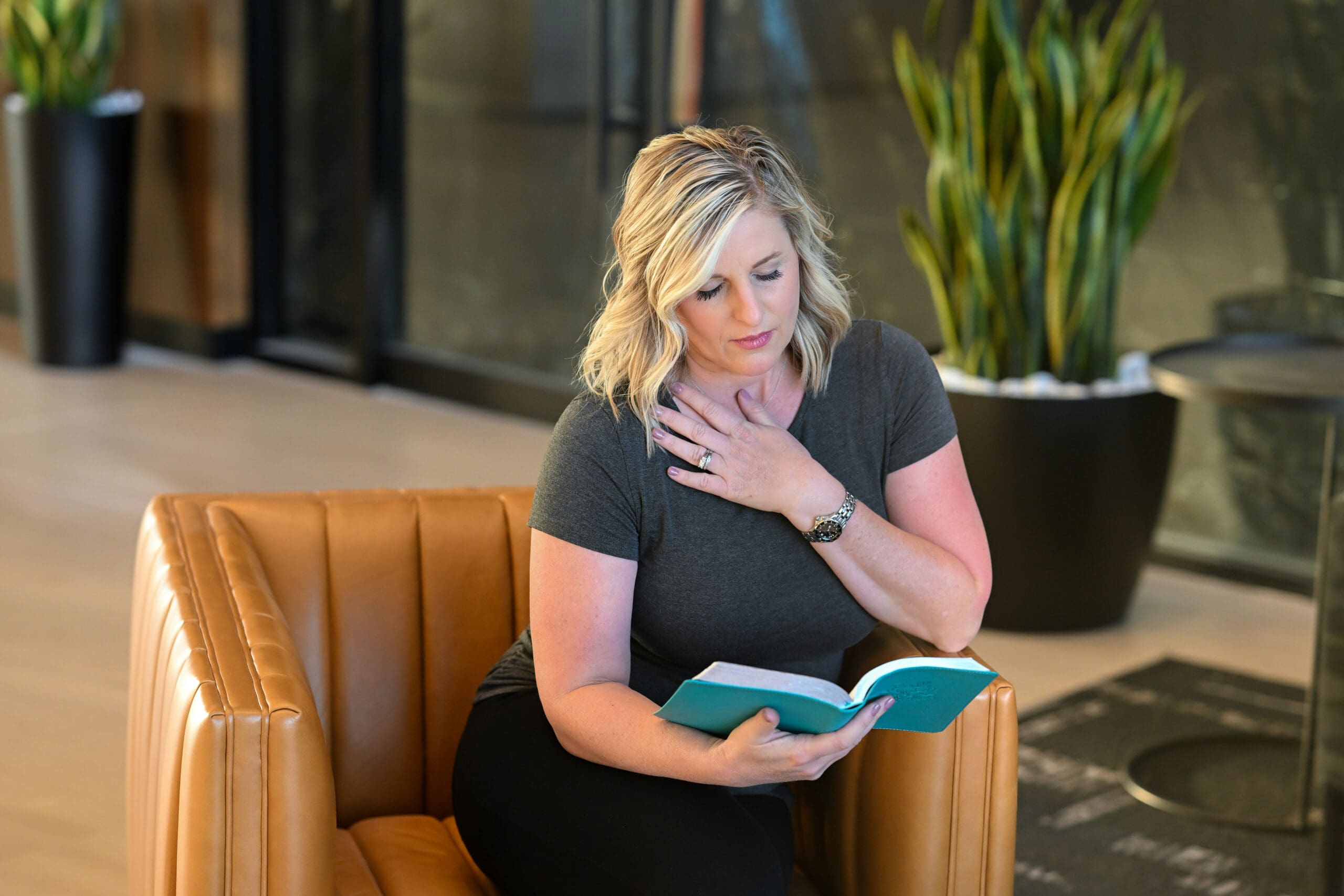 A woman with blonde wavy hair sits in a tan leather chair, holding a teal journal while placing her hand on her chest in a moment of reflection. She is wearing a dark gray top and black pants, with a peaceful yet emotional expression. The background features modern decor with green plants and a black table.