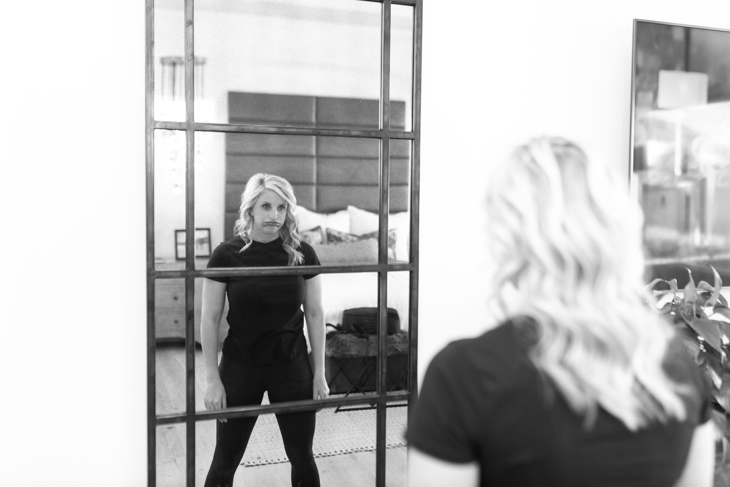 A woman with blonde hair and a disappointed expression stands in front of a large mirror, gazing at her reflection. The image conveys feelings of frustration and reflection, symbolizing the emotional toll of waiting and fertility struggles.