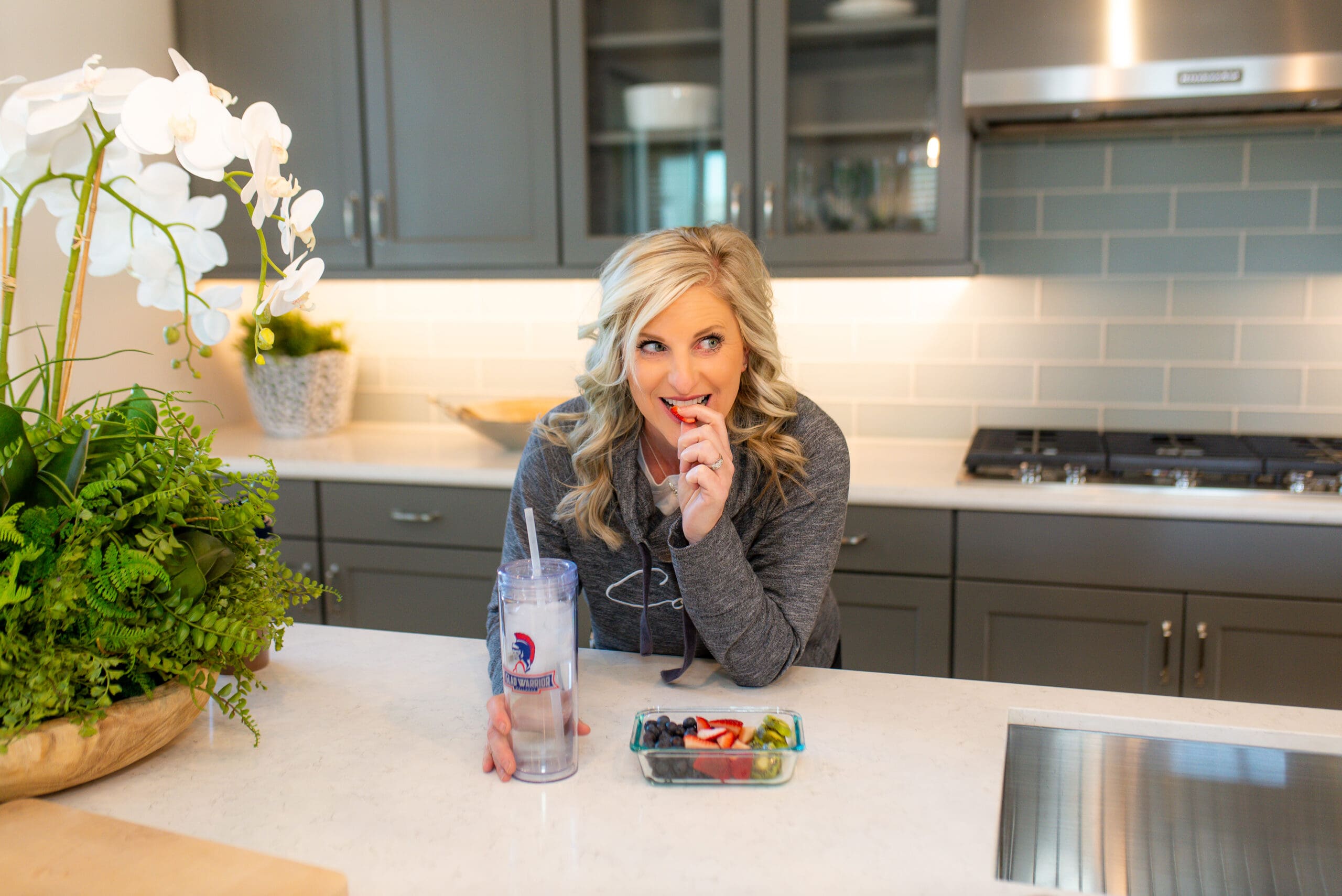 Woman enjoying a healthy snack in a bright kitchen, holding a cup of water and a bowl of fresh fruit while smiling.