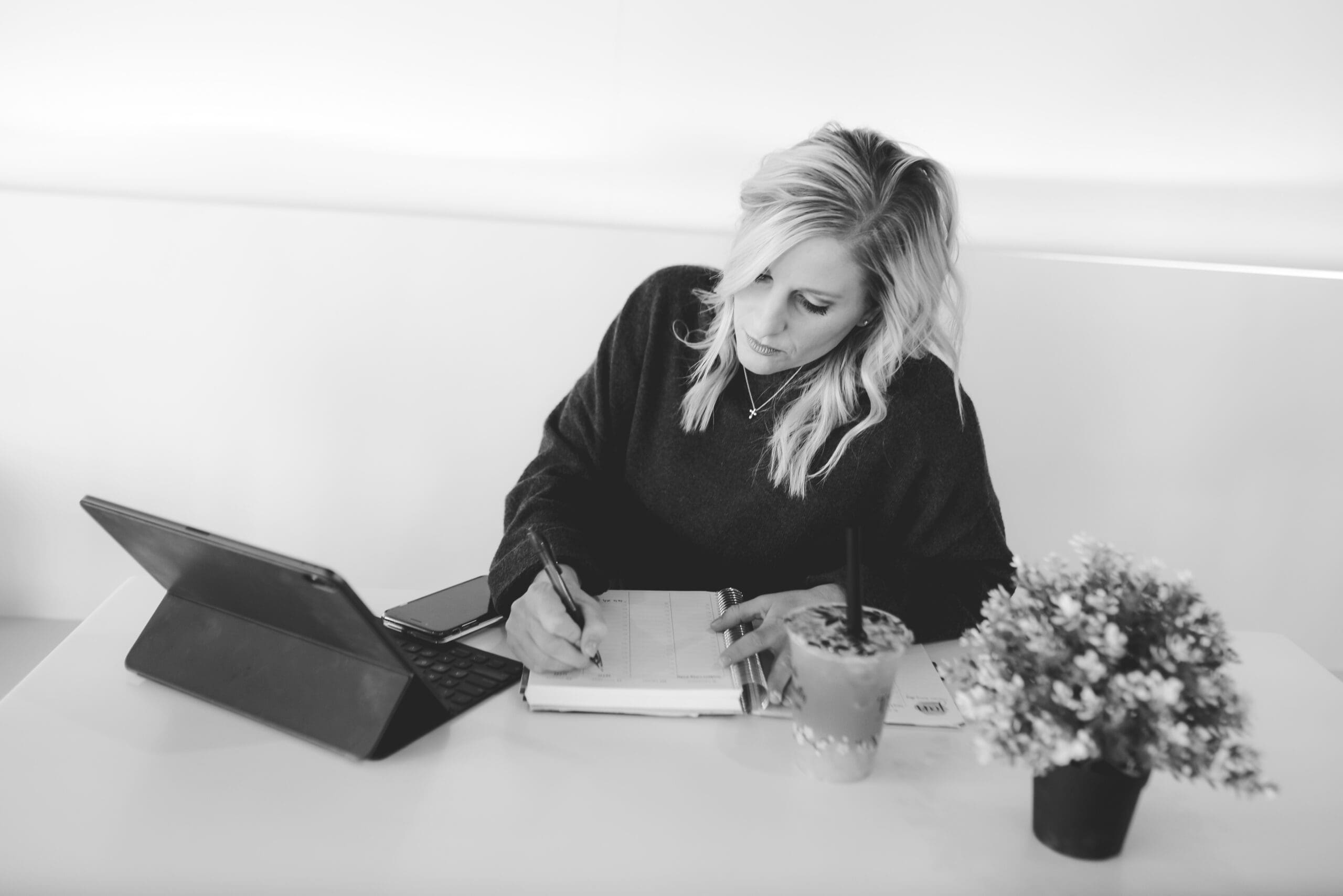 A woman with blonde hair sits at a white table, writing in her planner with a focused expression. A tablet, phone, and a smoothie with a straw are placed nearby, symbolizing intentional goal-setting and planning.