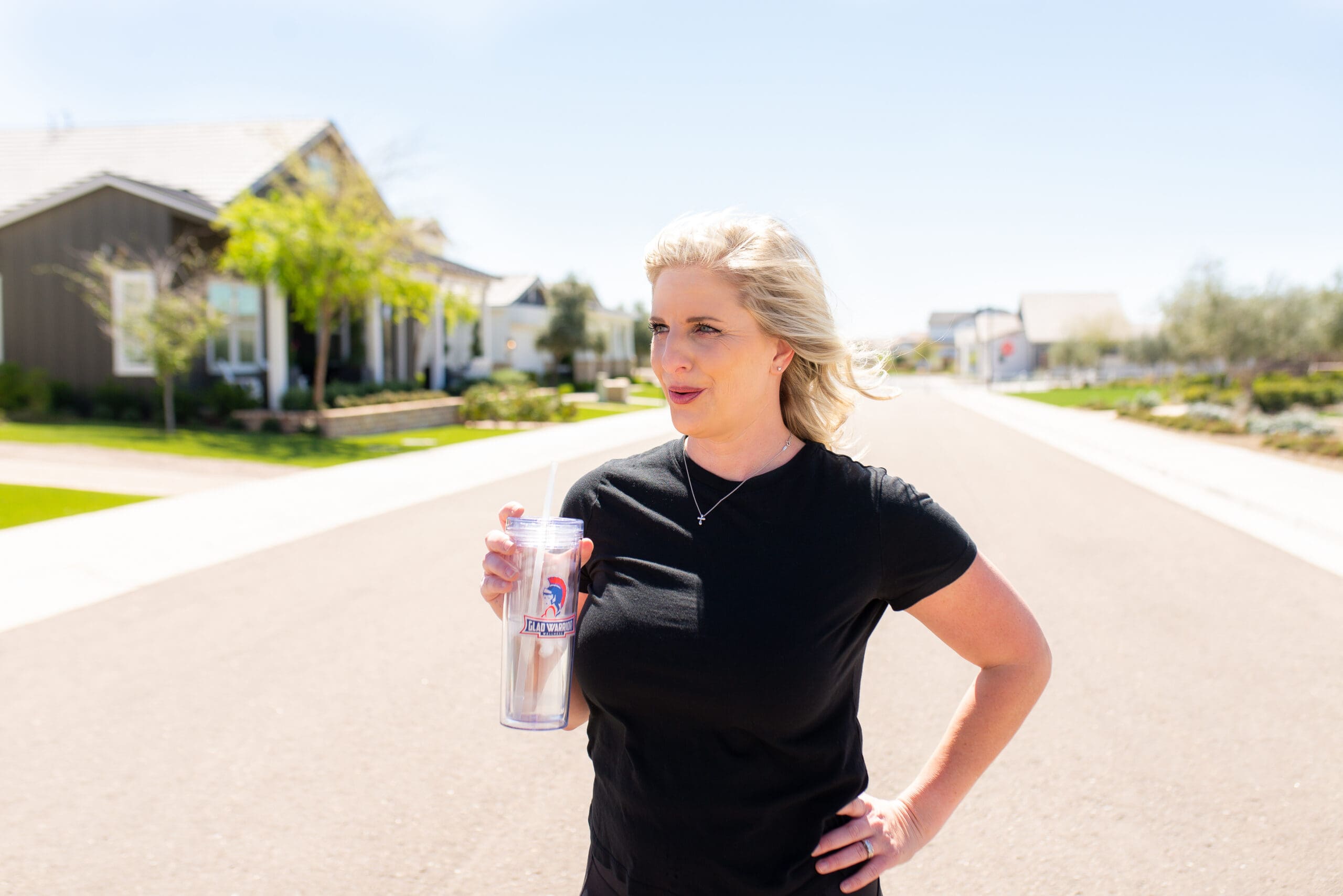 Woman in a black t-shirt standing on a sunny suburban street, holding a clear water tumbler, representing the importance of hydration, stress management, and healthy habits for fertility after vasectomy reversal.